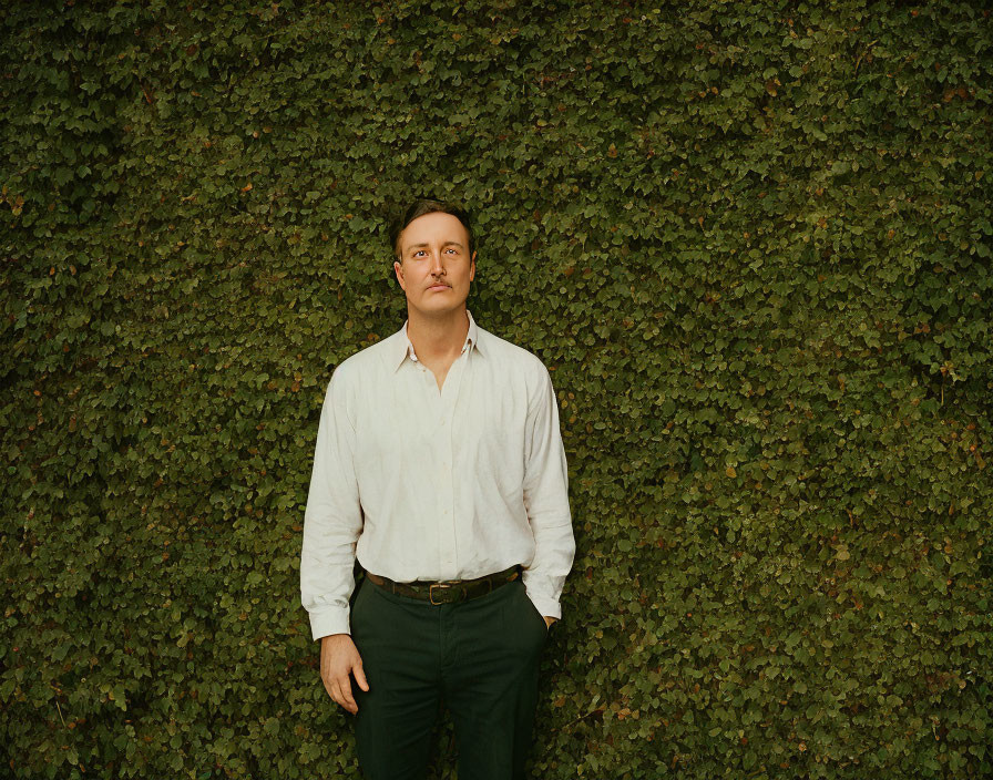 Man in white shirt and dark trousers standing before lush ivy wall