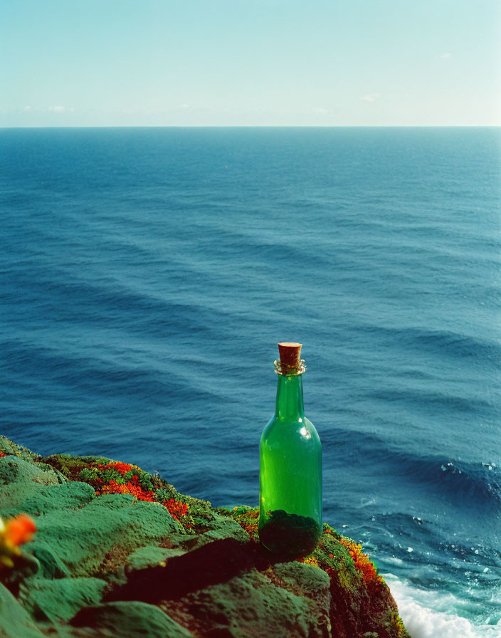 Green glass bottle with cork stopper on mossy coastal cliff overlooking serene ocean and sky.