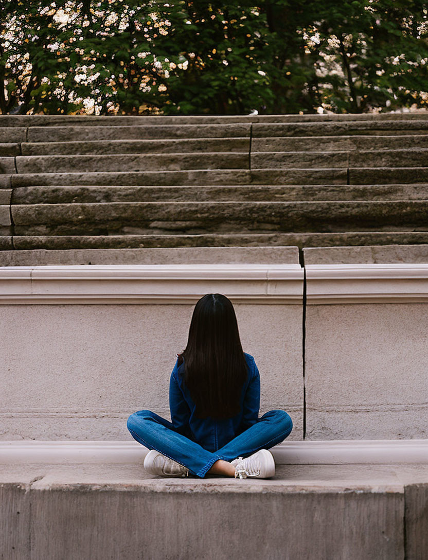 Long-haired person sitting on ledge near staircase and trees.