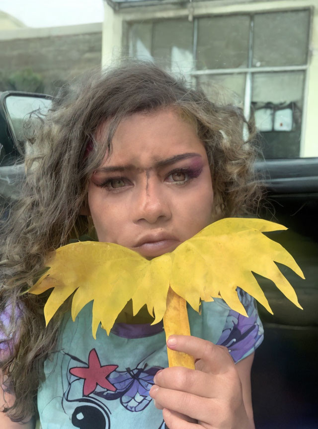 Curly Haired Person Holding Yellow Paper Mustache Outdoors