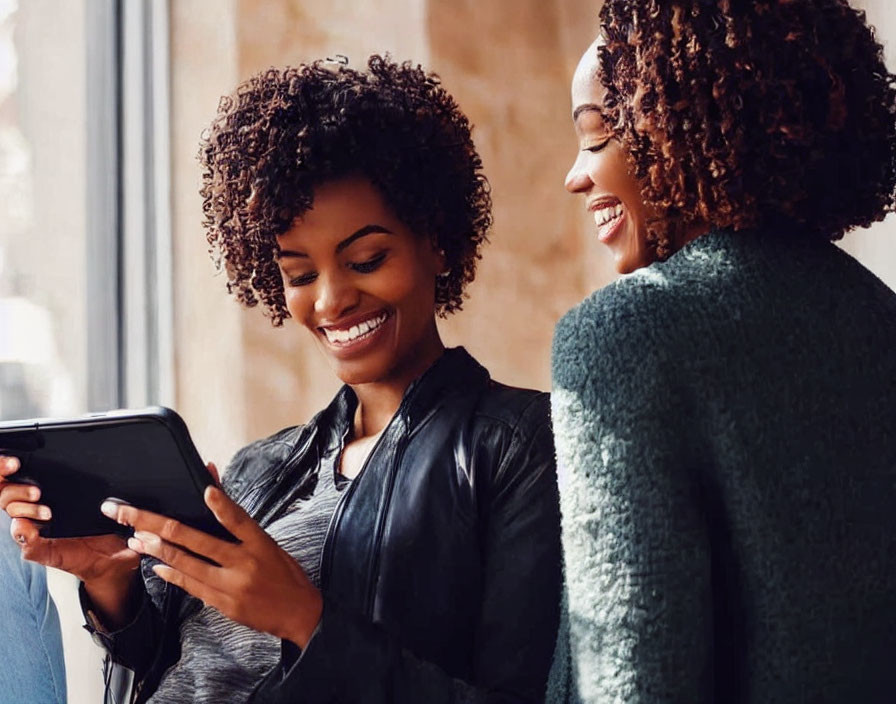 Two women with curly hair smiling at tablet screen