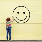 Woman in striped shirt next to smiley face drawing on yellow wall