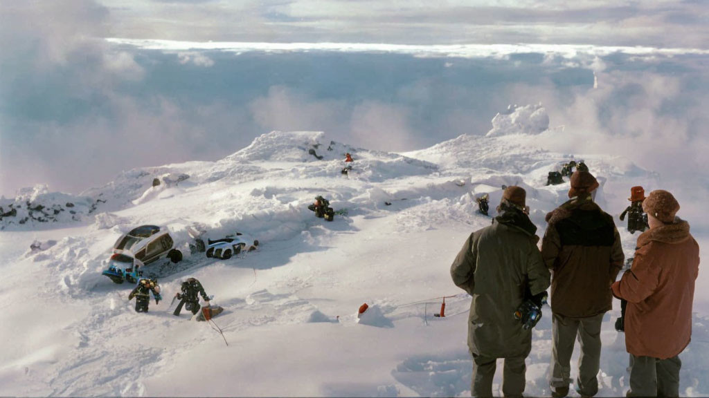 Mountain peak covered in snow with people and vehicles, surrounded by clouds on a sunny day