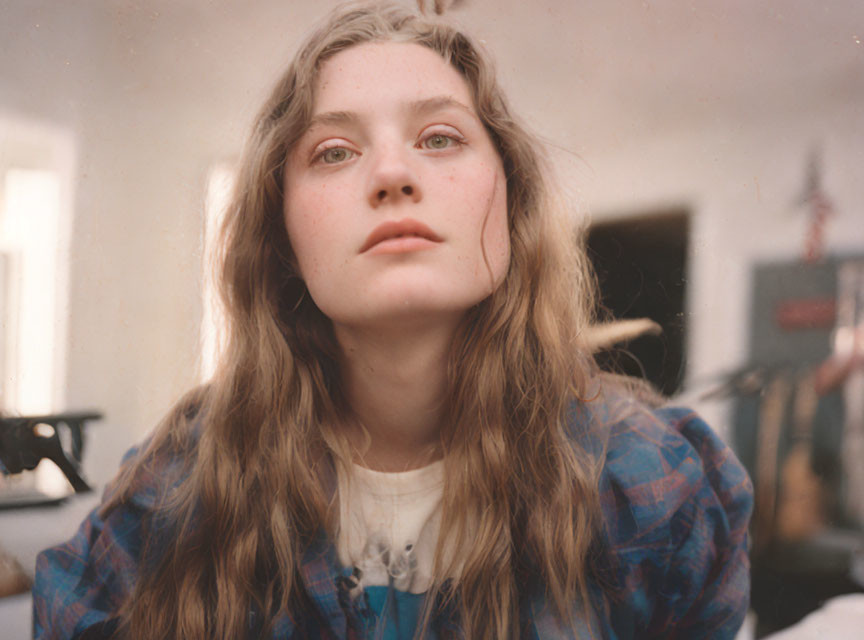 Young woman with long wavy hair in checkered shirt gazes upwards in soft indoor light