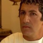 Middle-aged man with dark hair, mustache, and stubble in white shirt in room with beige