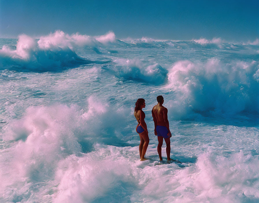 Two individuals on a beach near foamy waves under clear sky