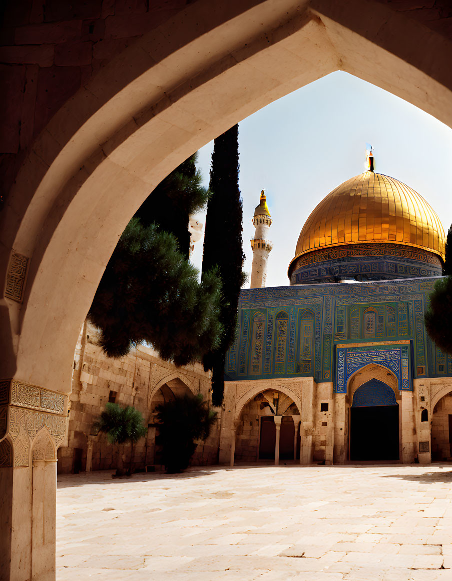 Golden dome and blue facade of Dome of the Rock framed by arch, with trees and clear sky.