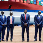 Five men in suits posing by a red ship under a clear blue sky