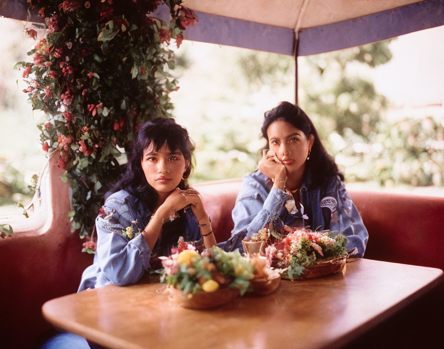 Two women in denim outfits enjoying food at a table outdoors with lush plant background