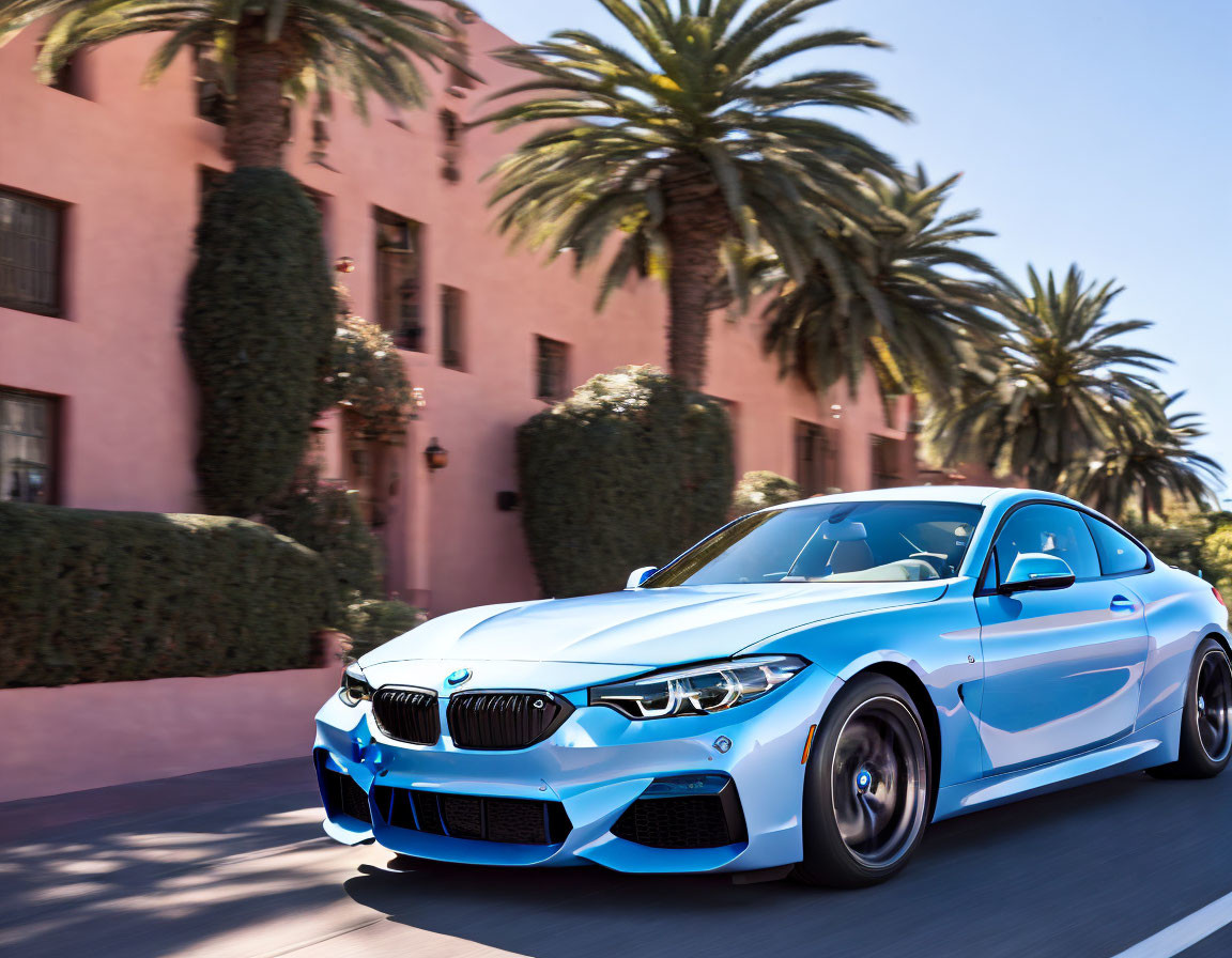 Blue BMW coupe driving past pink building with shrubbery under clear sky