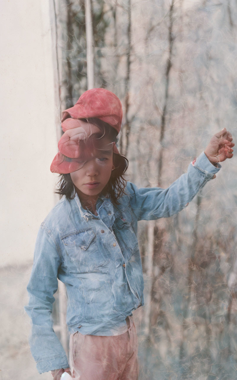 Child in denim jacket and pink hat by frosted window, looking pensive