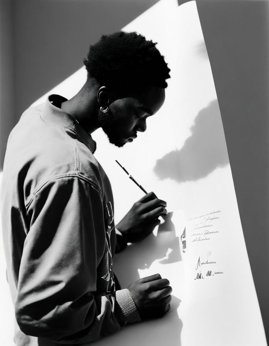 Man in denim jacket writing on large paper with pen under sunlight.