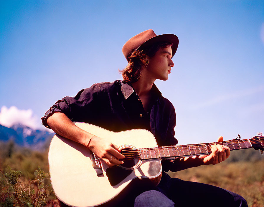 Person in Brown Hat Playing Acoustic Guitar Outdoors with Mountain Background