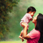 Mother lifting smiling toddler under tree with sunlight.