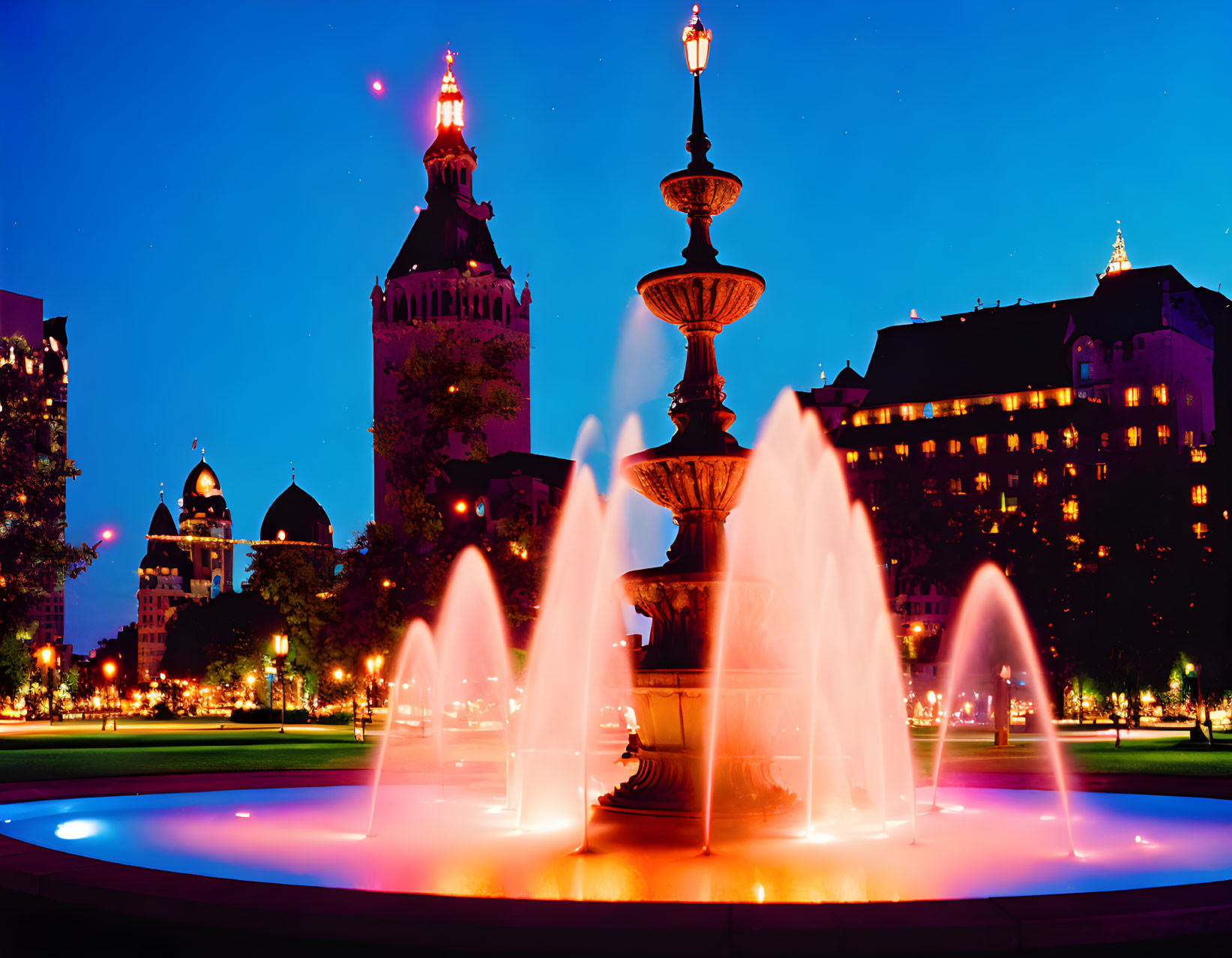 Twilight scene: illuminated fountain, arcing water jets, historical building, clear evening sky