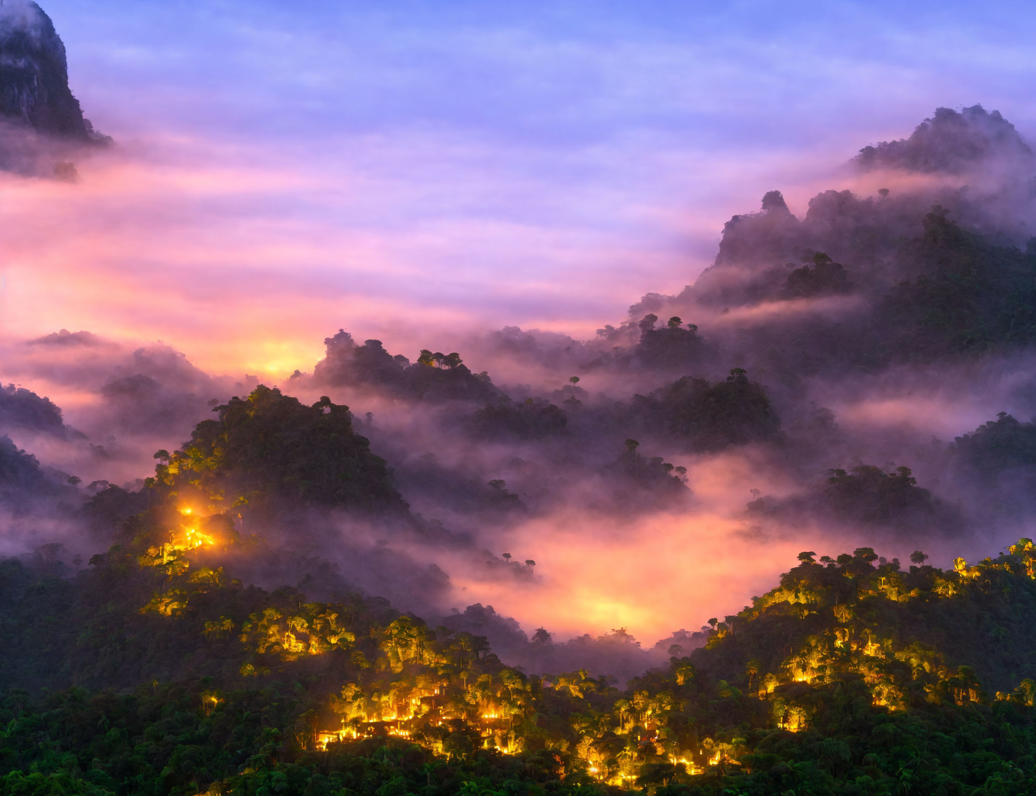 Vivid sunset over misty jungle with mountain silhouettes & warm village lights.