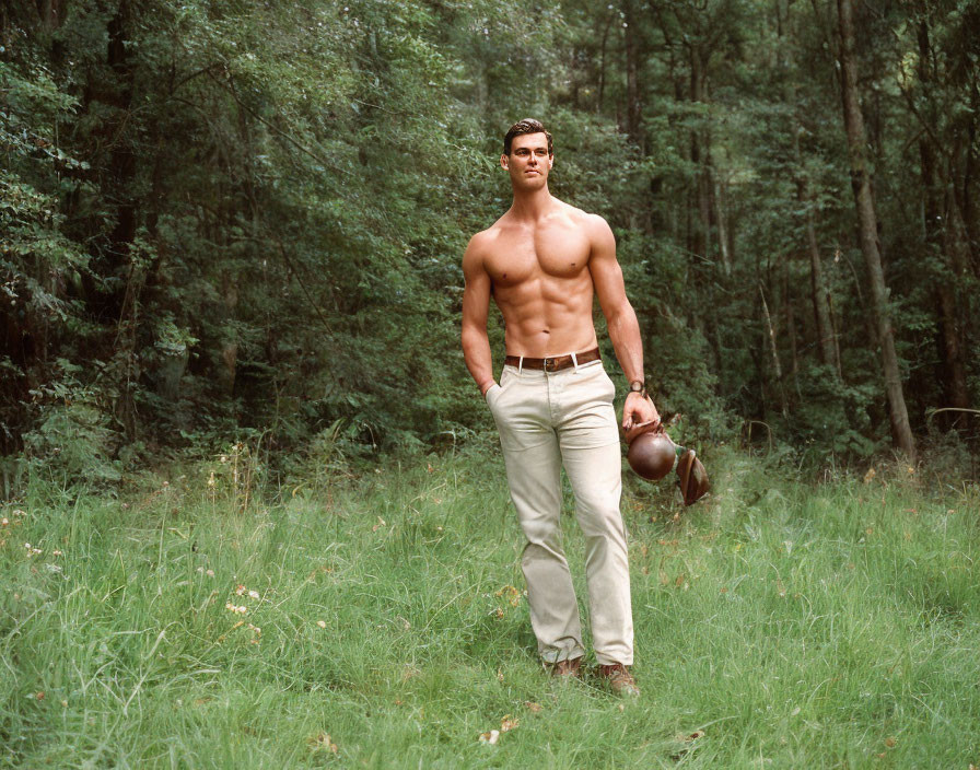 Muscular man in beige trousers holds football helmet in forest