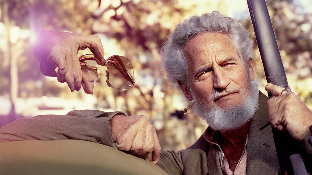 Elderly man with white beard holding sunglasses by car with blurred trees.