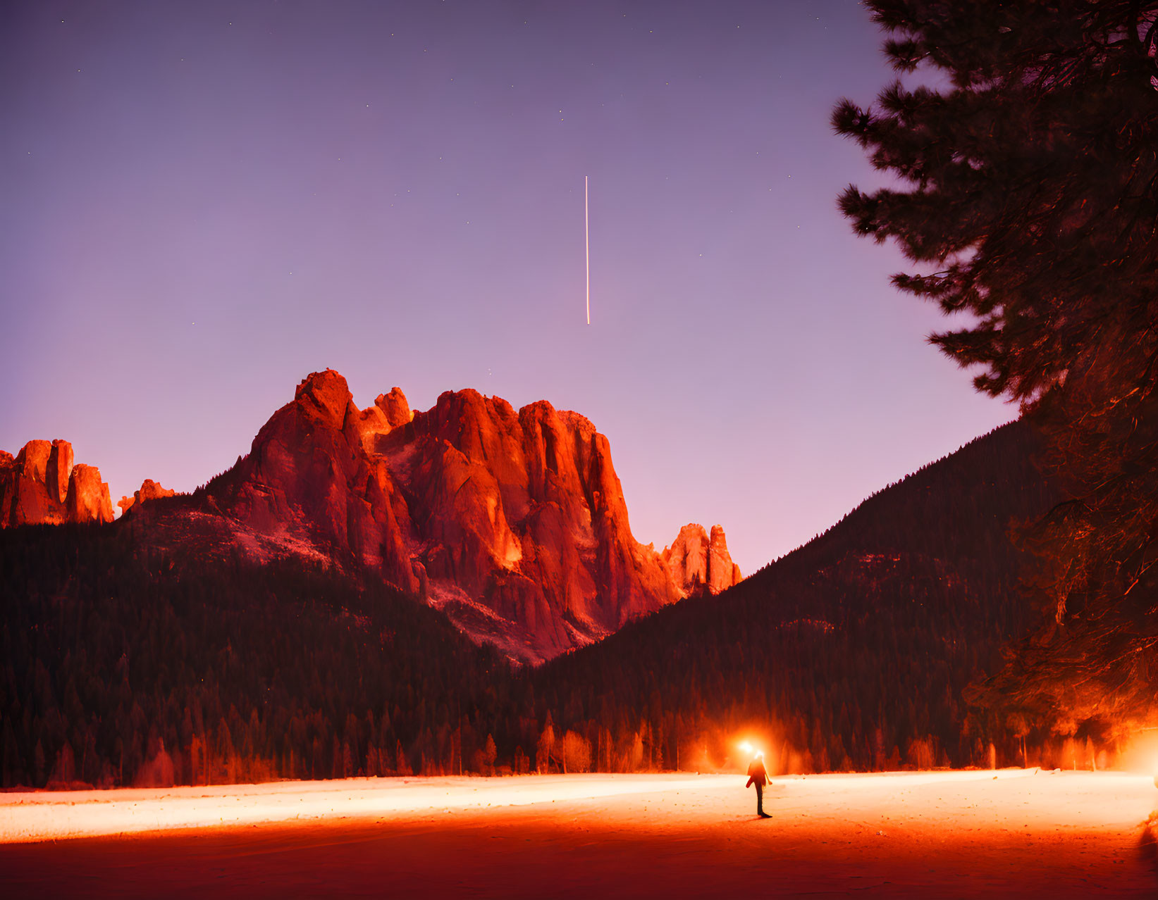 Person under starry sky with shooting star above rugged mountains and campfire glow