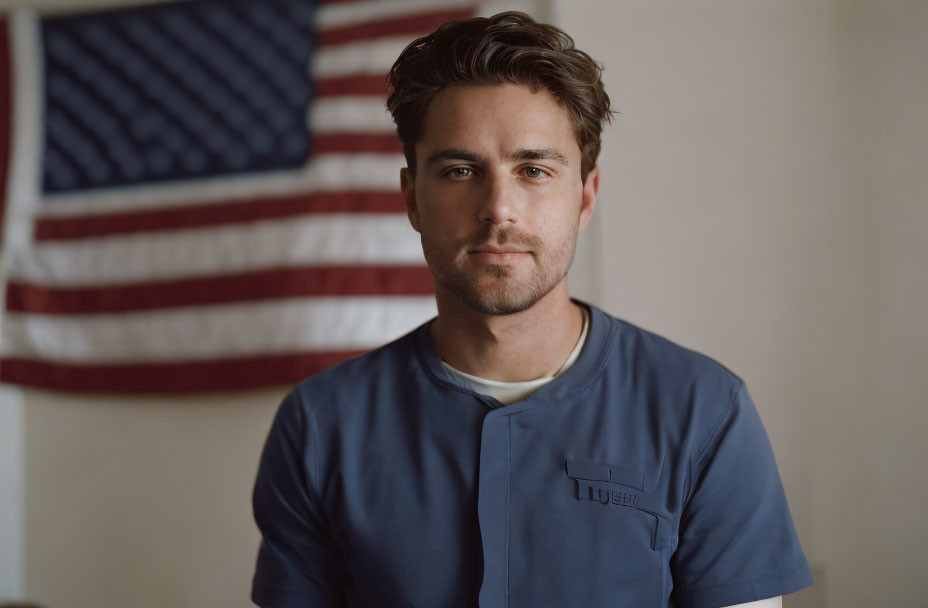 Male healthcare worker in blue scrubs poses in front of American flag.