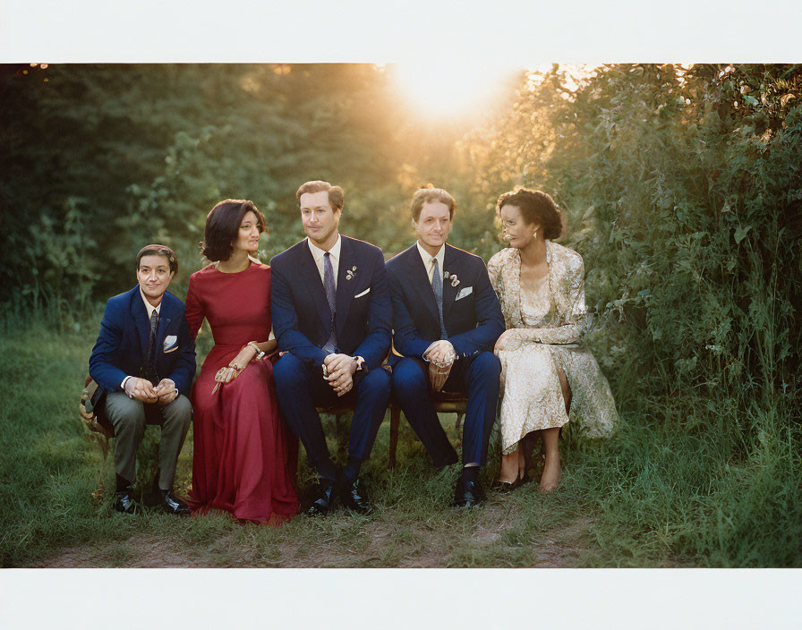 Five adults in elegant attire sitting on bench outdoors under sunlight.