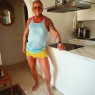 Gray-bearded man with tattoos in sunglasses, tank top, and yellow shorts in modern kitchen