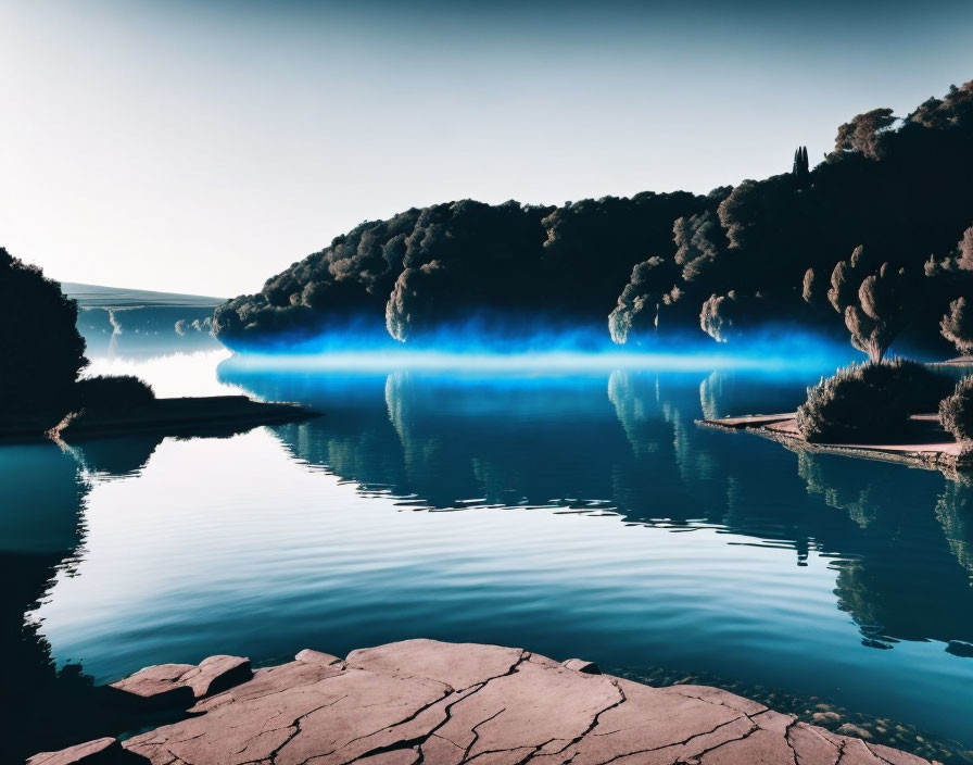 Tranquil lake with mist, forested hills, clear sky, and rocky shore