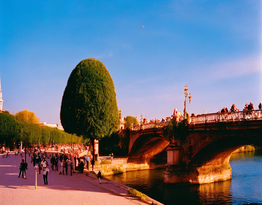 Bustling riverside walkway with bridge statues and clear sky