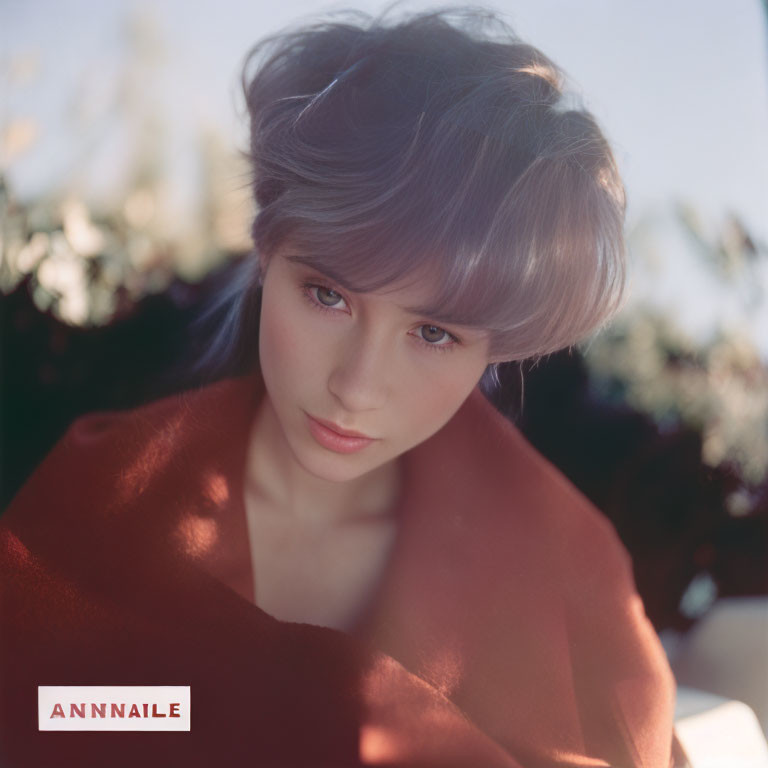 Portrait of woman with blue hair and striking eyes in orange garment with "ANNAILE" text.