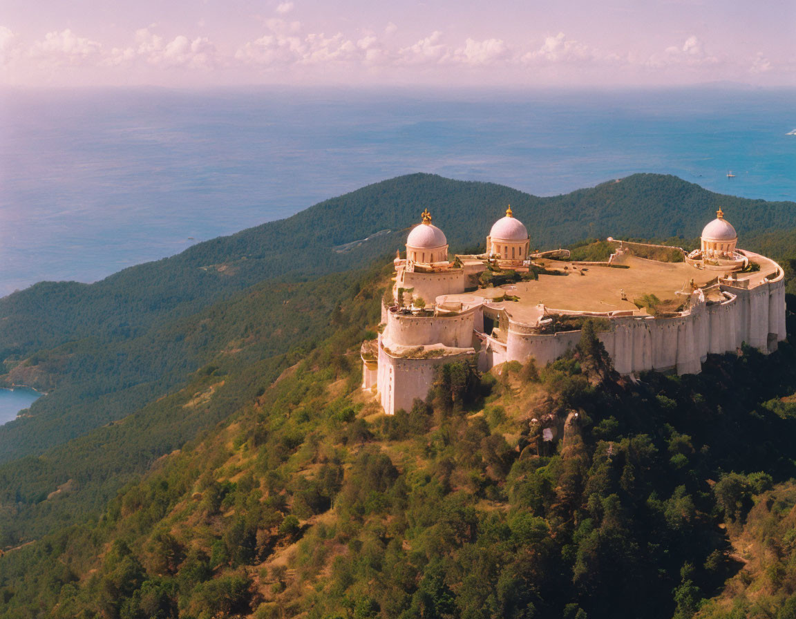 Fortress with domed structures on hilltop amidst forests and coastline.