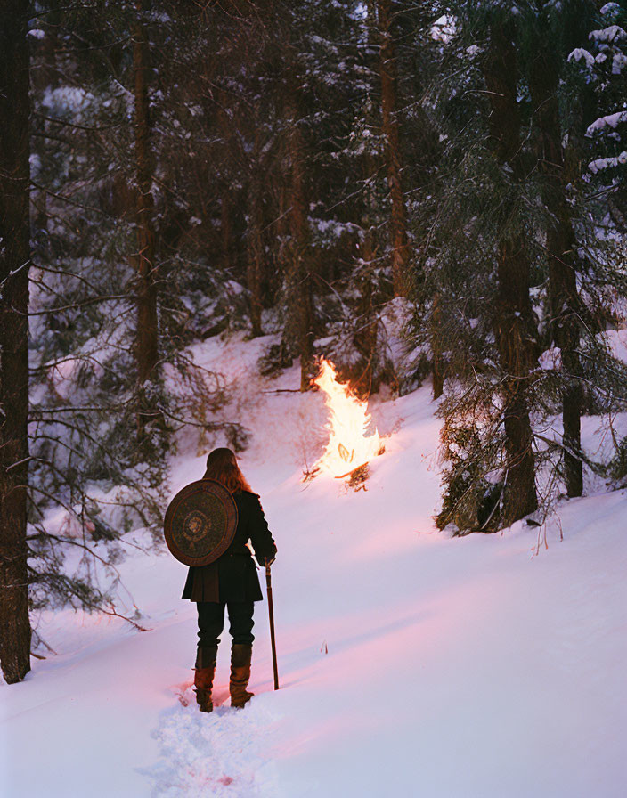 Person in winter attire with shield and staff in snowy forest near small fire.