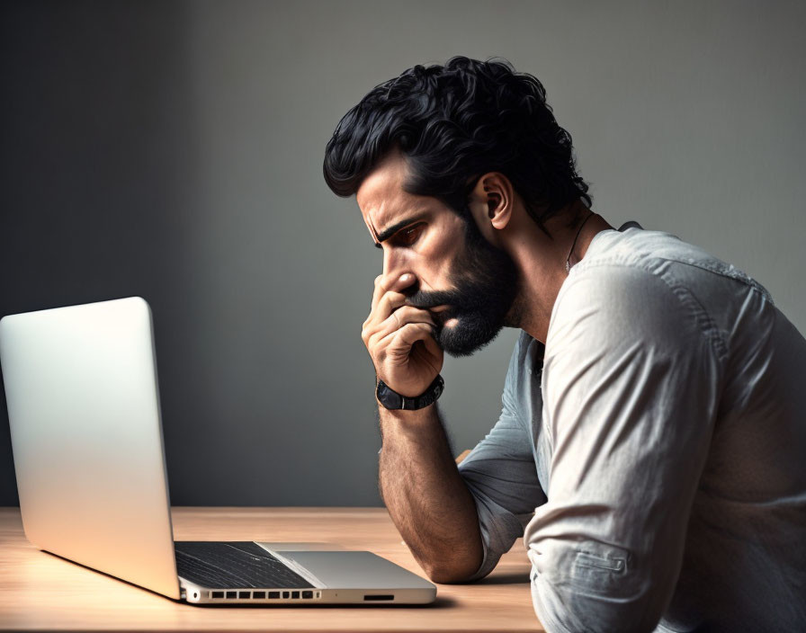 Bearded man at desk gazes at laptop thoughtfully