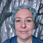 Blue-haired person in makeup and outfit against snowy forest.