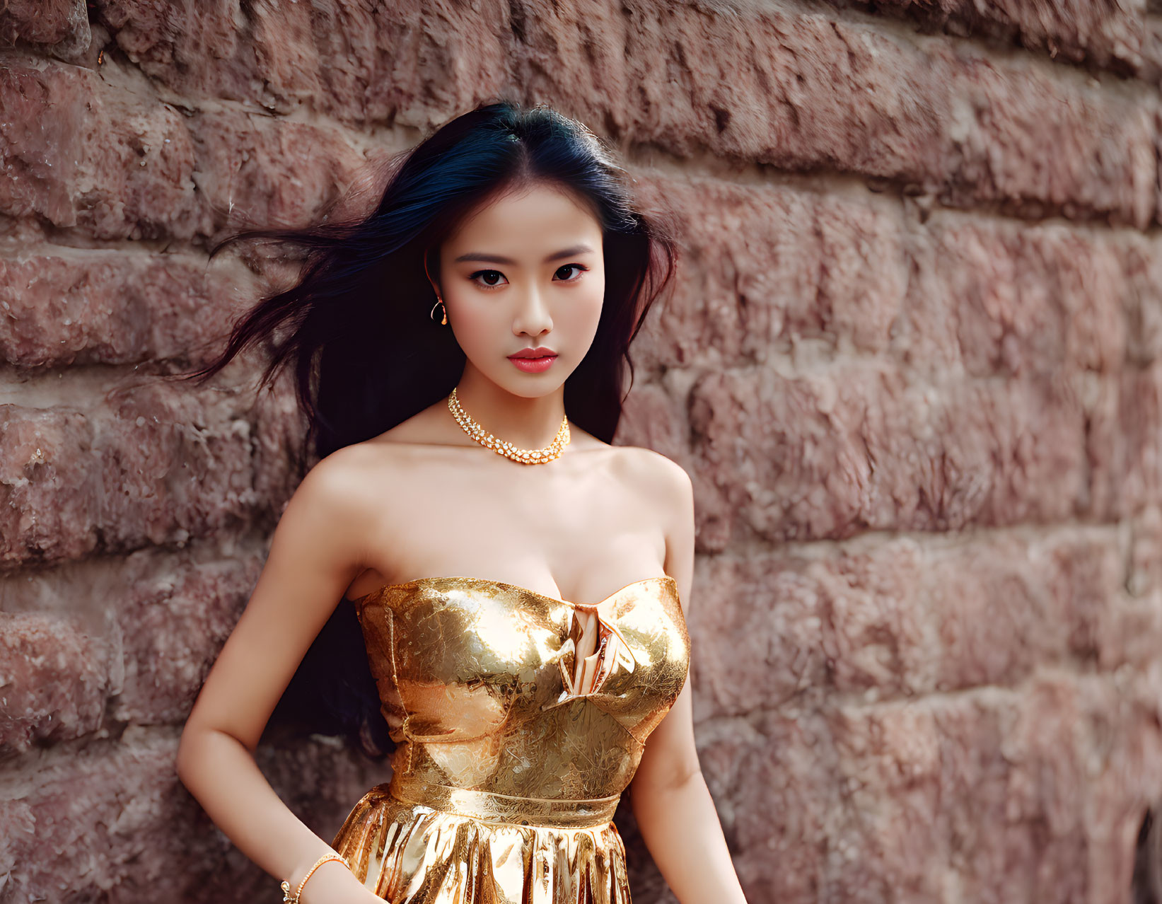 Dark-haired woman in golden strapless dress against brick wall