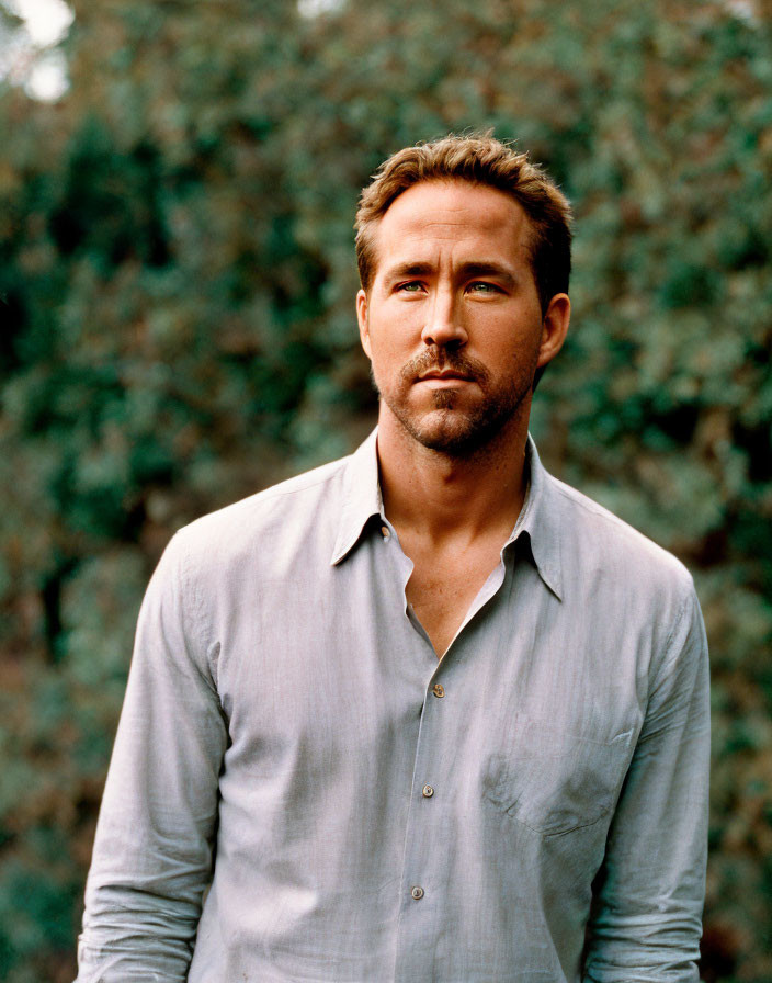 Man with stubble in gray shirt against green leafy backdrop