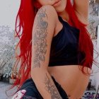 Vibrant red-haired woman with floral tattoos in black top and denim shorts