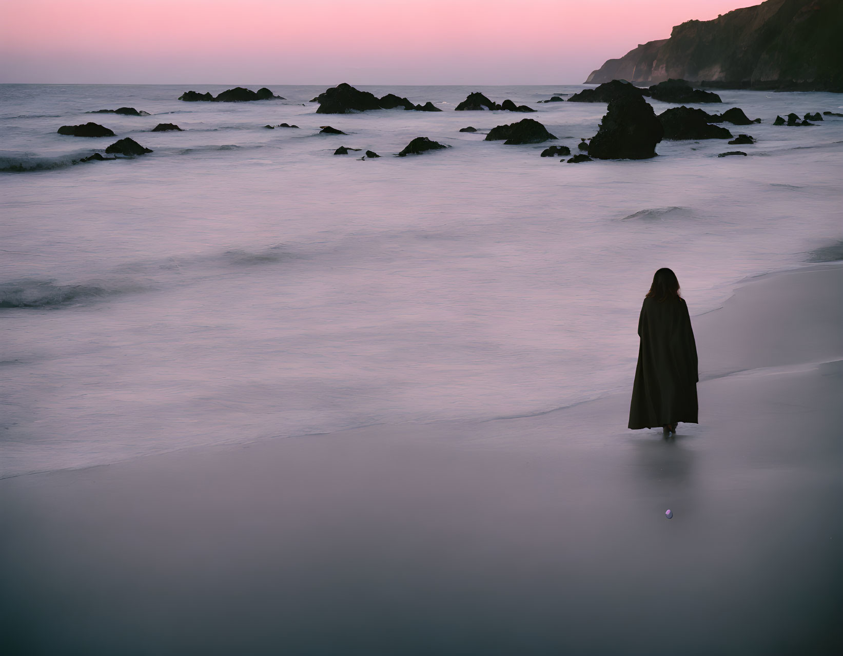 Cloaked figure on twilight beach with scattered rocks and hazy shoreline.