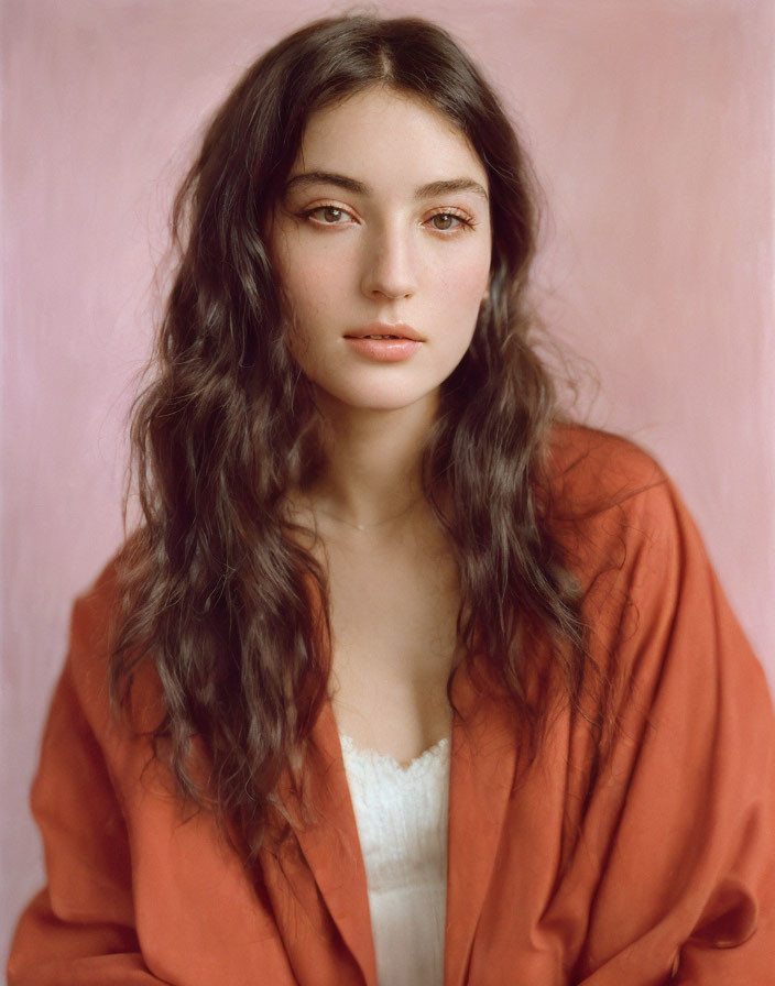 Portrait of woman with long wavy hair in white top and orange garment on pastel pink background