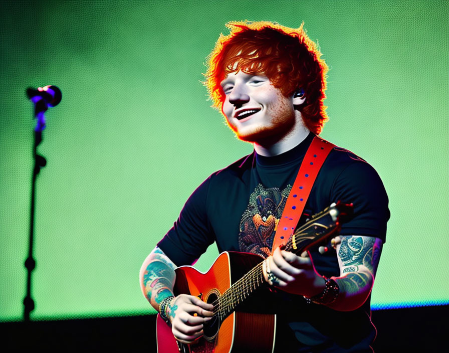Red-haired male musician playing acoustic guitar with tattoos, smiling on stage.