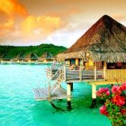 Sunset overwater bungalow with balcony, turquoise sea, flowers, and palm trees