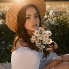 Woman in straw hat with flowers, sitting in daisy field.