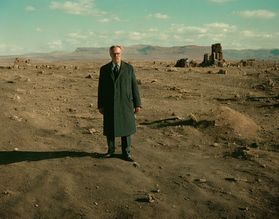 Man in dark coat in barren landscape with ruins and distant mountains