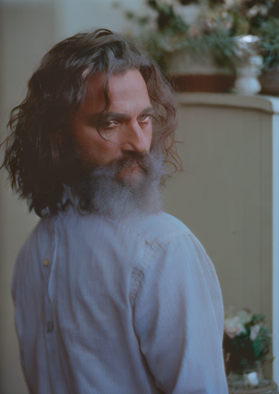 Bearded man with long curly hair in light blue shirt gazes pensively