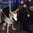 Woman in dark dress with floral headpiece near white unicorn in mystical forest.