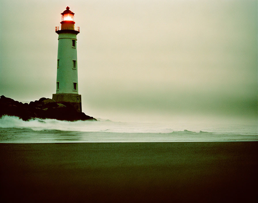 White lighthouse with red top on rocky shore by misty sea and green sky