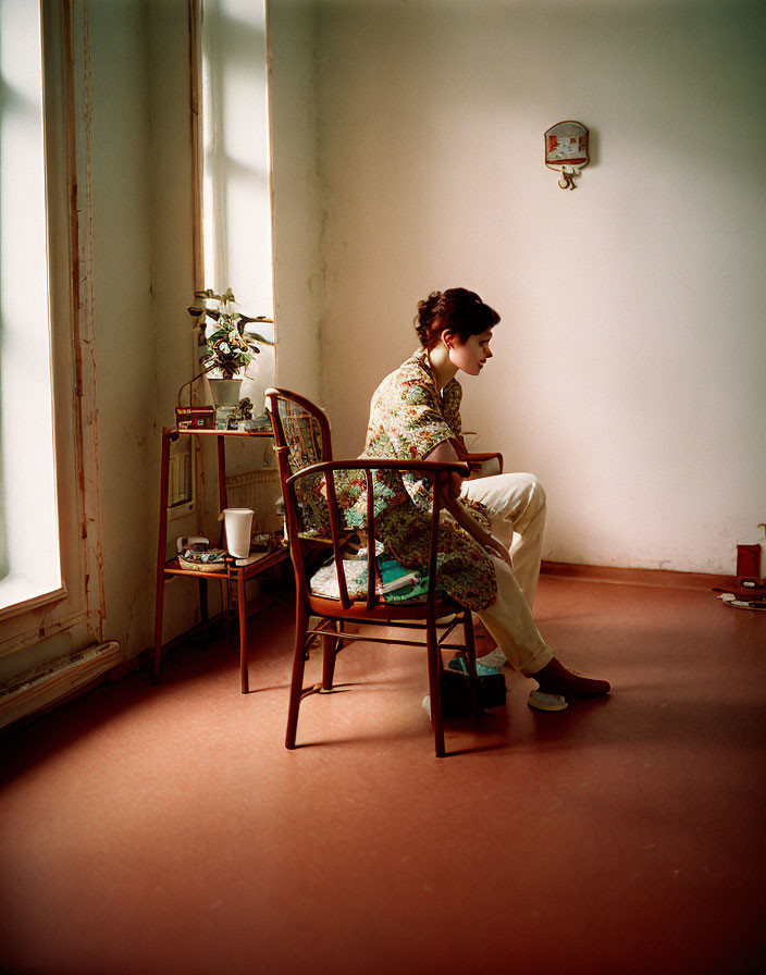 Person in floral outfit seated on wooden chair in sunny room with plant and thermometer.