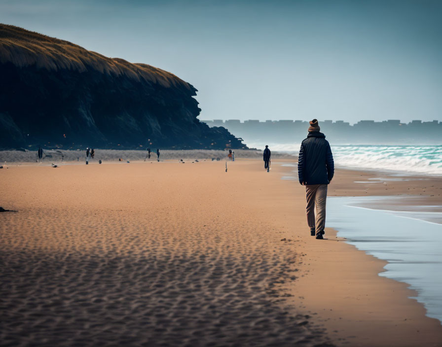 Person in jacket and hat walking on sandy beach with sea and cliff.