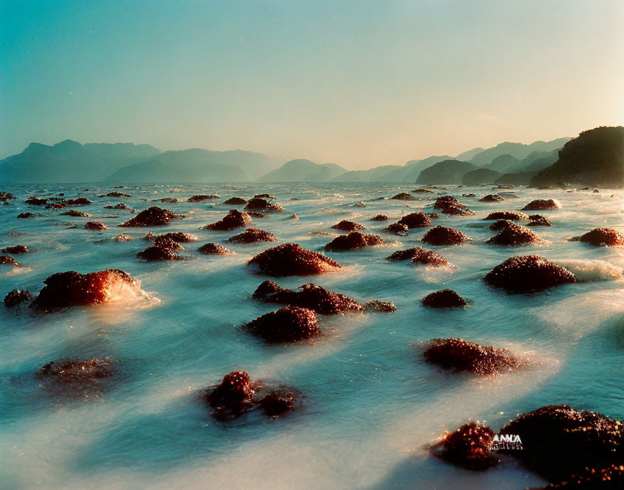 Tranquil Coastal Sunset with Rocks, Waves, and Misty Mountains
