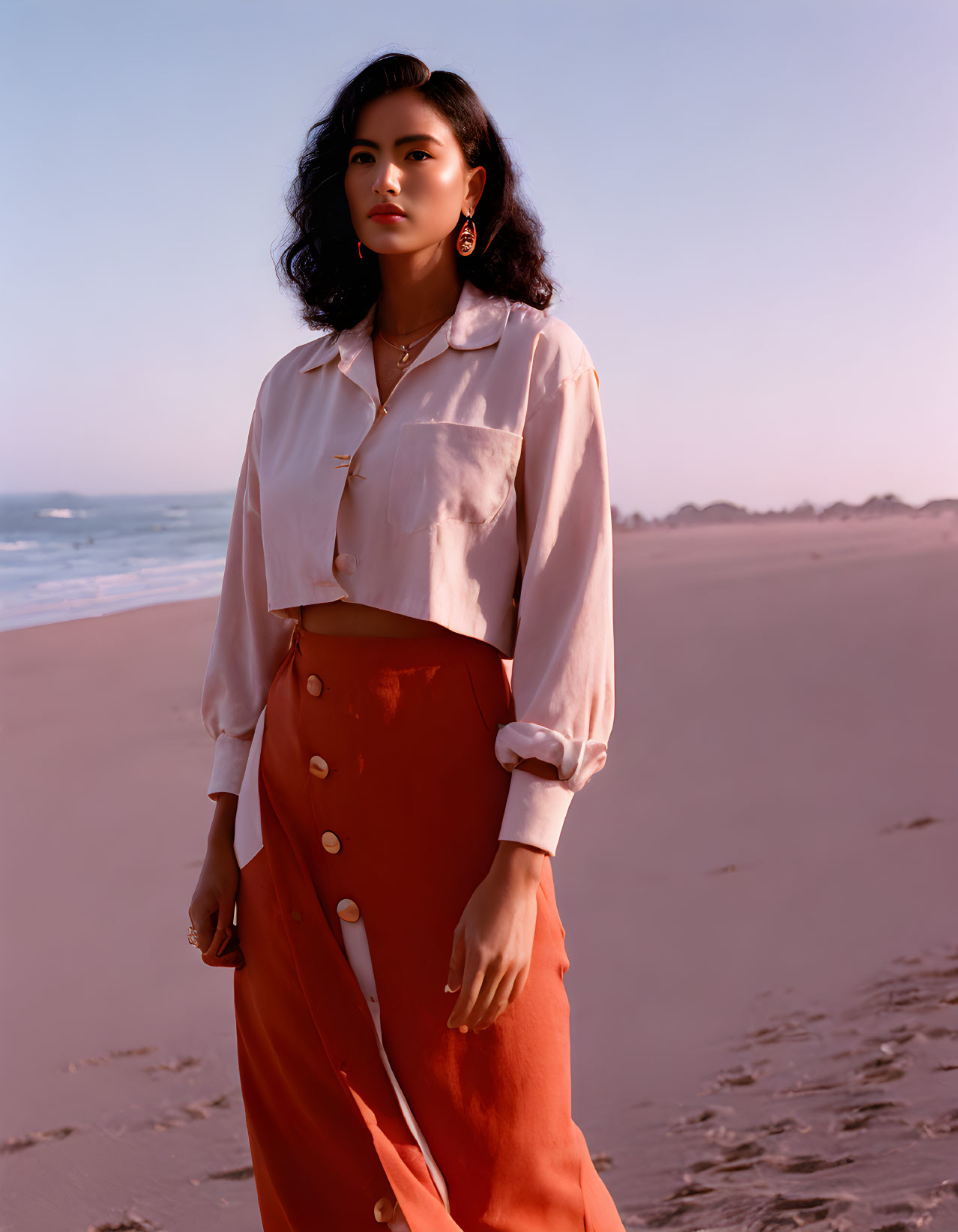 Woman in white blouse and terracotta skirt on beach with hand on hip, gold jewelry.