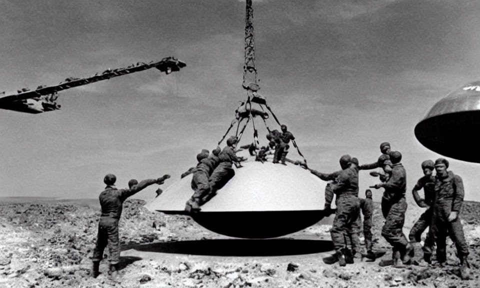 Military men lift UFO-like object with crane in desert.